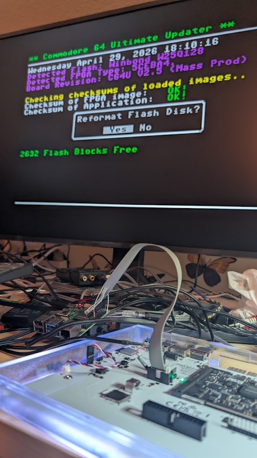 Photo of an HDMI screen showing the Commodore 64 Ultimate Updater menu with 'Detected FPGA type', 'Board Revision: C64U V2.5 (Mass Prod)' and a 'Reformat Flash Disk?' yes/no prompt. Below the screen, the open Commodore 64 Ultimate is visible on a desk with a JTAG ribbon cable connected to its P5 header.